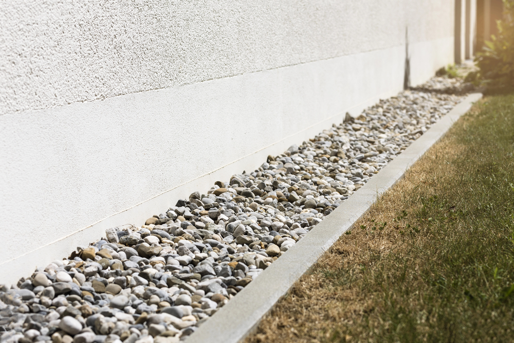 A narrow strip of decorative rocks lines the base of a white exterior wall, bordered by concrete edging and adjacent to a patch of dry grass.