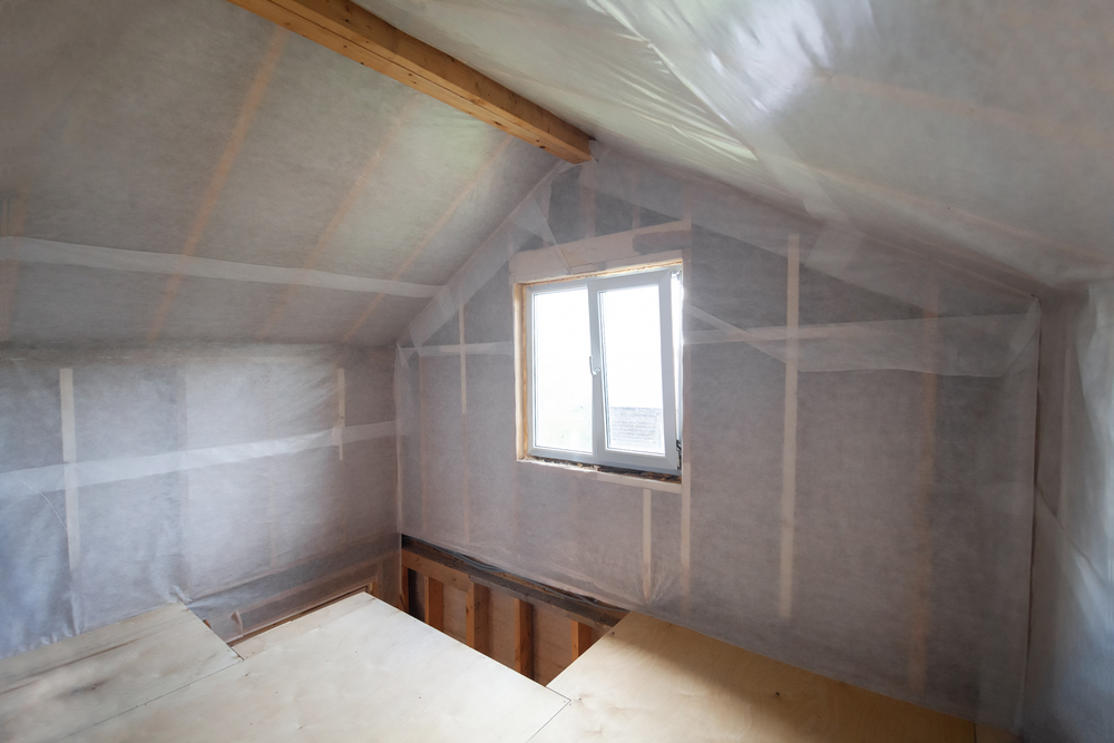 An unfinished attic room with exposed wooden beams, insulation, and a small window letting in natural light.
